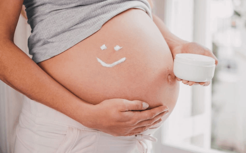 pregnant lady is holding her belly while from another hand she is holding stretch mark cream