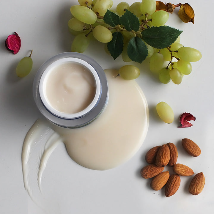 An open jar of cream surrounded by almonds, green grapes, and rose petals, with a soft cream spill creating a natural, nourishing aesthetic.